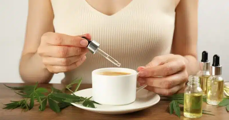 A person adding a drop of CBD or THC tincture into a cup of tea, surrounded by hemp leaves and bottles of tincture.