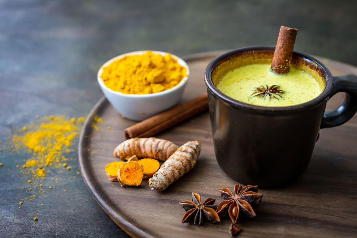 A warm cup of golden milk with turmeric, cinnamon, and star anise, served on a wooden tray with fresh turmeric root and a bowl of ground turmeric.