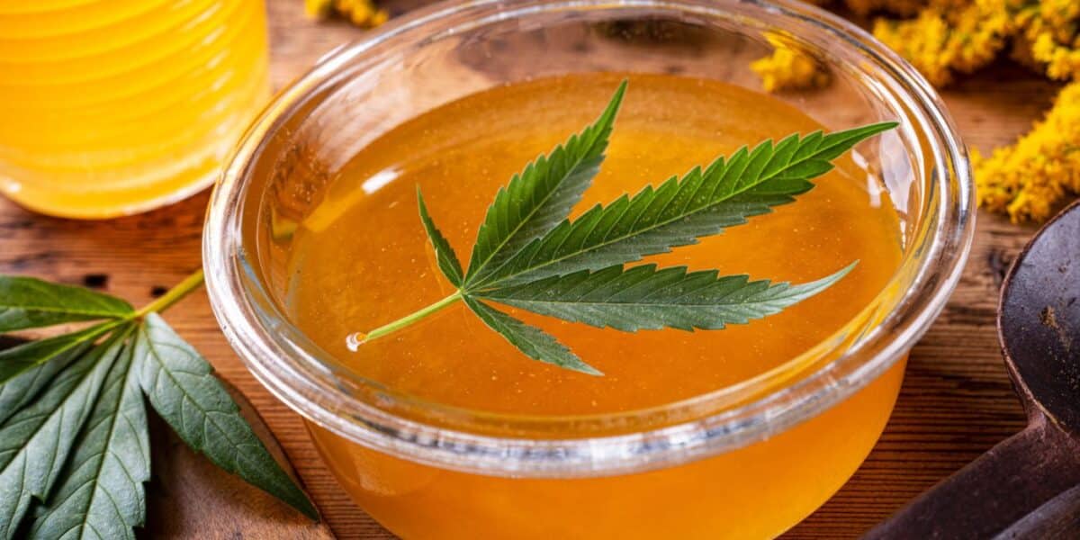 CBD honey in a clear glass bowl with a cannabis leaf on top, surrounded by organic honey jars, cannabis leaves, and yellow flowers on a wooden table