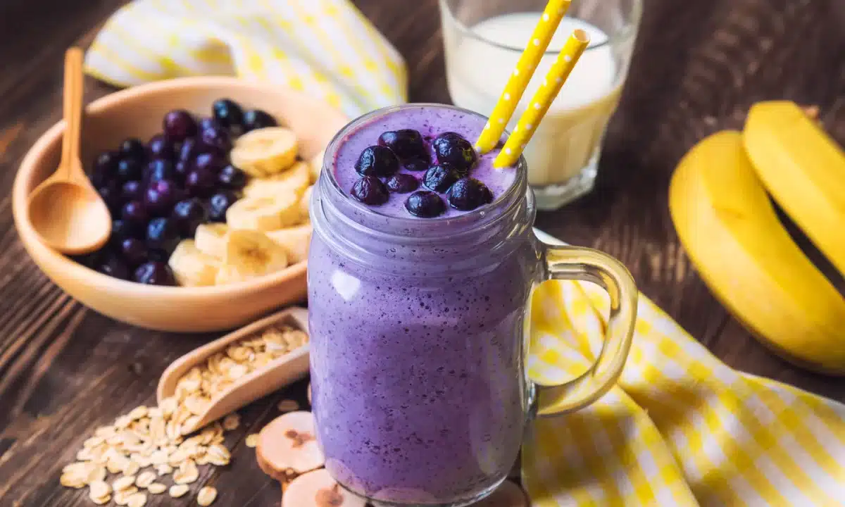 A blueberry banana smoothie in a mason jar with yellow polka dot straws, surrounded by fresh ingredients like bananas, oats, and blueberries.