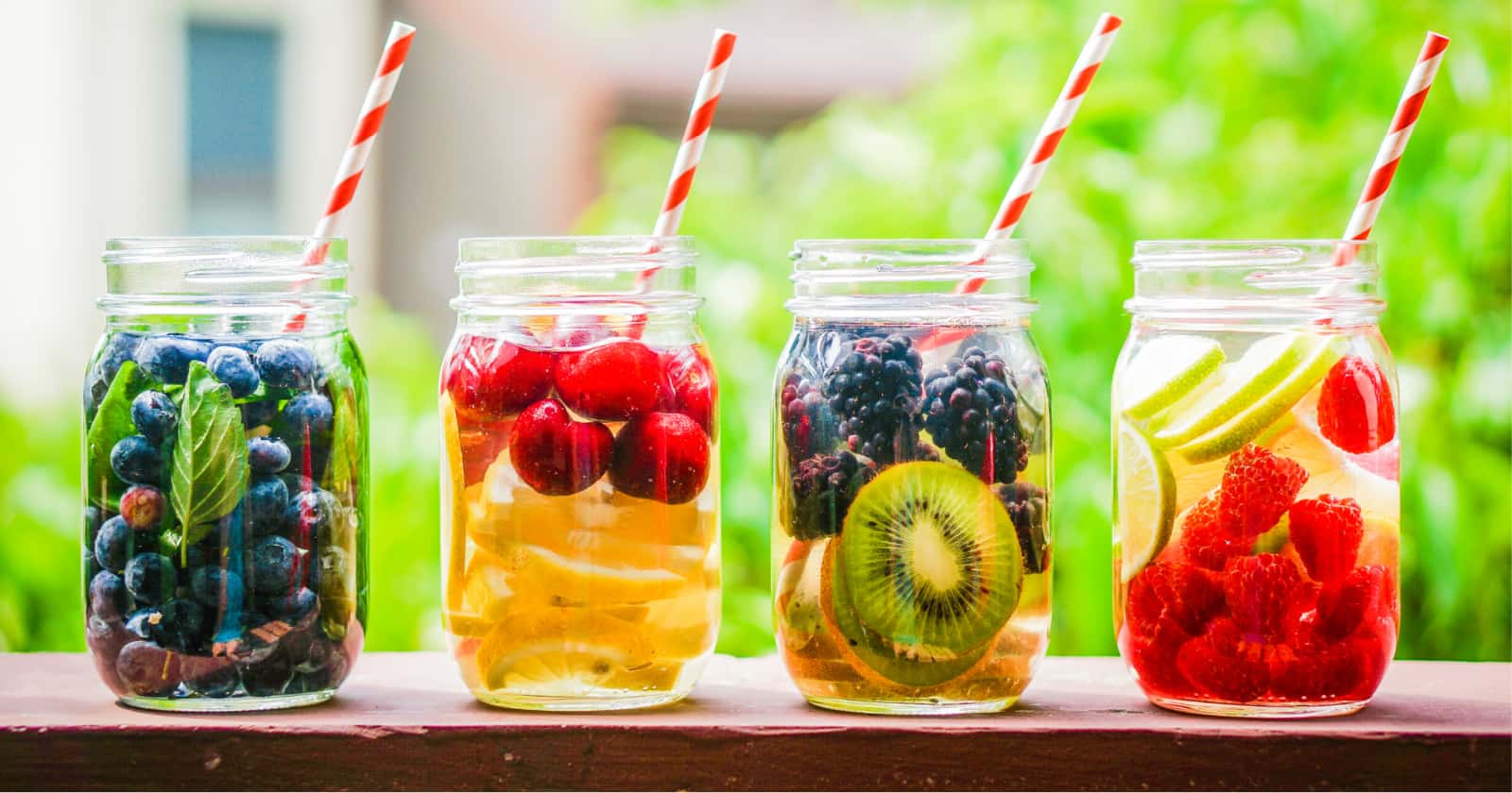 Four mason jars filled with fruit-infused water, featuring berries, citrus, and kiwi, with striped paper straws.