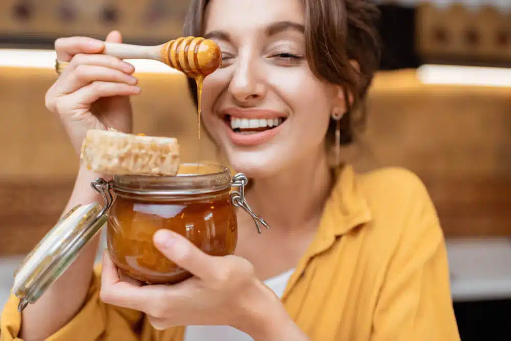 Smiling woman drizzling organic honey from a wooden dipper into a glass jar, showcasing natural honey benefits for CBD honey products from TheCannaCompany.com