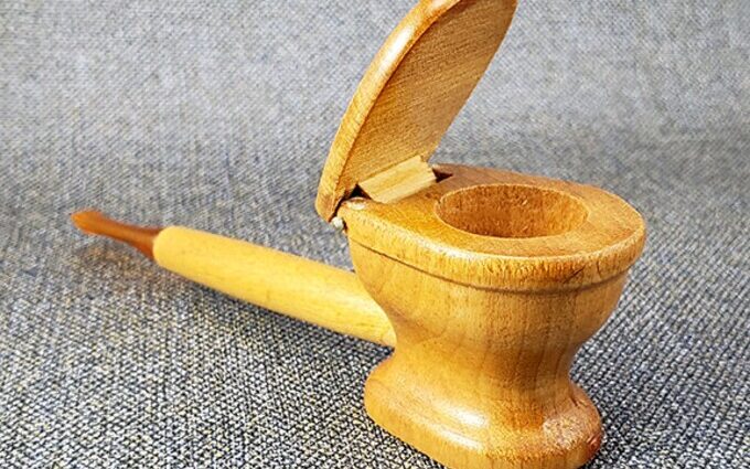 Handcrafted wooden novelty smoking pipe designed as a miniature toilet with an open lid and a long stem, placed on a textured gray fabric background.