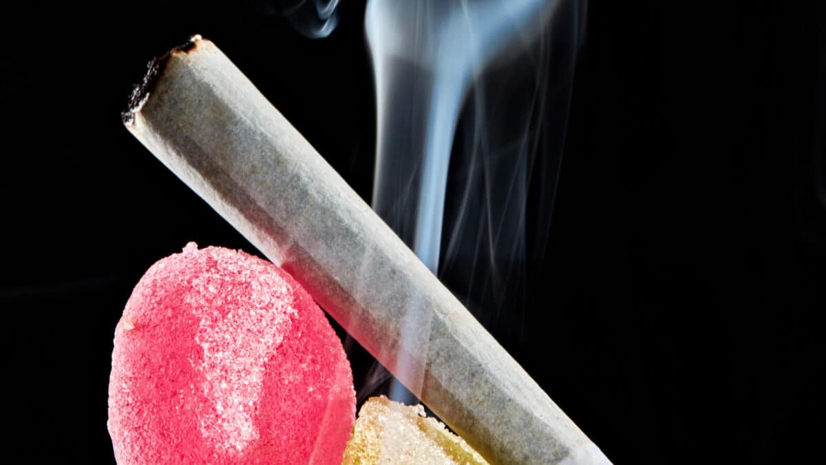 Close-up of a burning cannabis joint beside colorful THC-infused edibles with smoke rising on a black background.