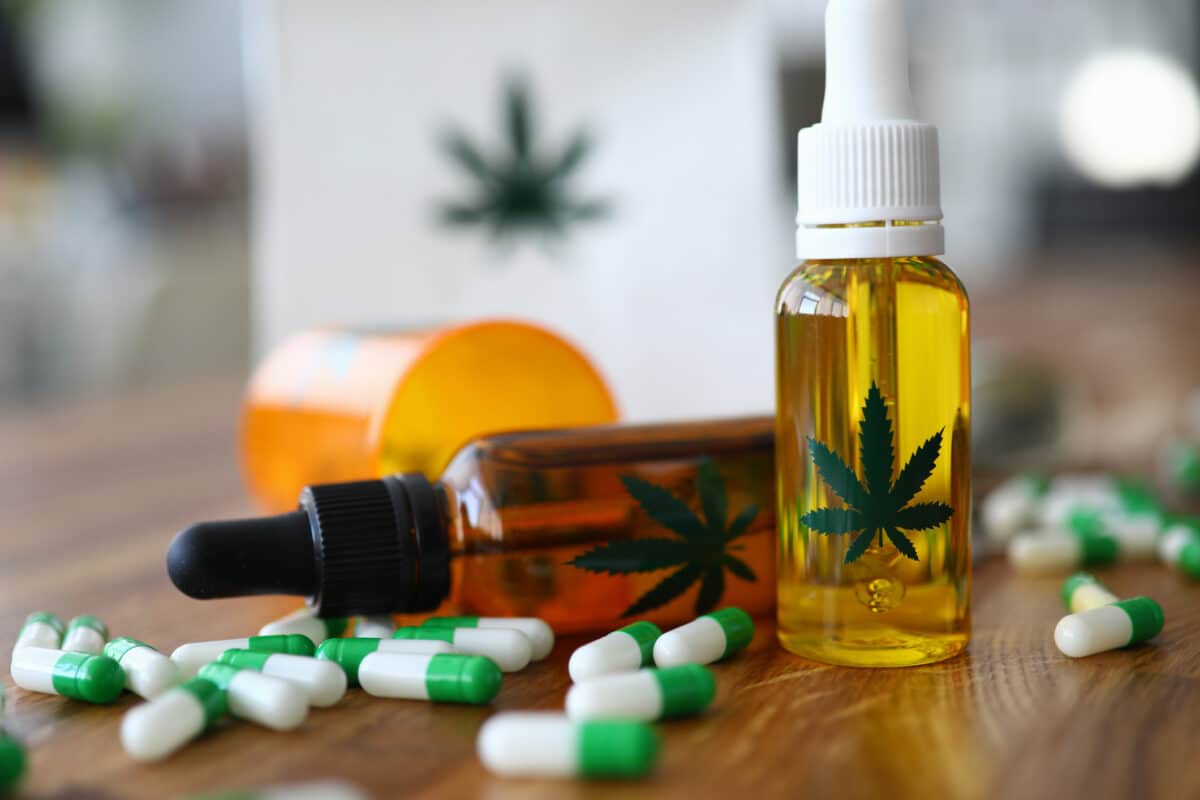Two CBD oil tincture bottles with cannabis leaf labels alongside scattered green and white pharmaceutical capsules and an orange prescription bottle on a wooden surface, illustrating the contrast between plant-based cannabidiol and conventional pharmaceutical drugs