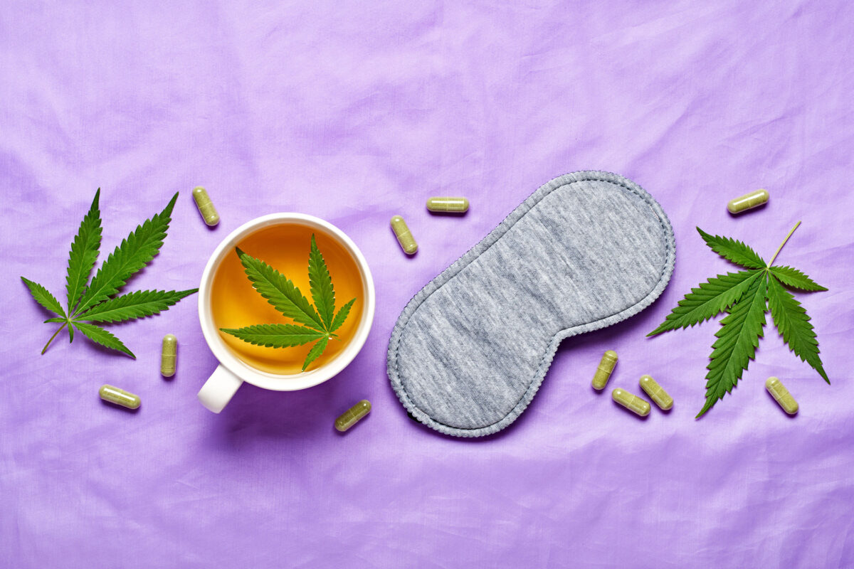 Flat lay of a white mug of golden cannabis tea with a cannabis leaf, a grey sleep mask, and scattered green CBD capsules on a purple background, representing cannabidiol as a natural sleep aid