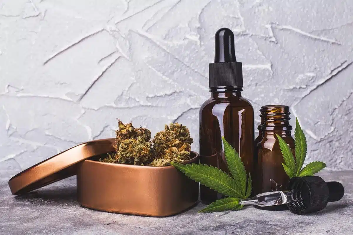 Two amber CBD oil tincture dropper bottles alongside a copper tin filled with cannabis flower buds and a fresh cannabis leaf on a textured grey surface, representing different forms of CBD wellness products